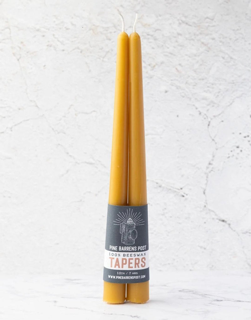 A pair of amber-orange beeswax taper candles joined by a navy blue paper band with brand name "Pine Barrens Post" and illustration.