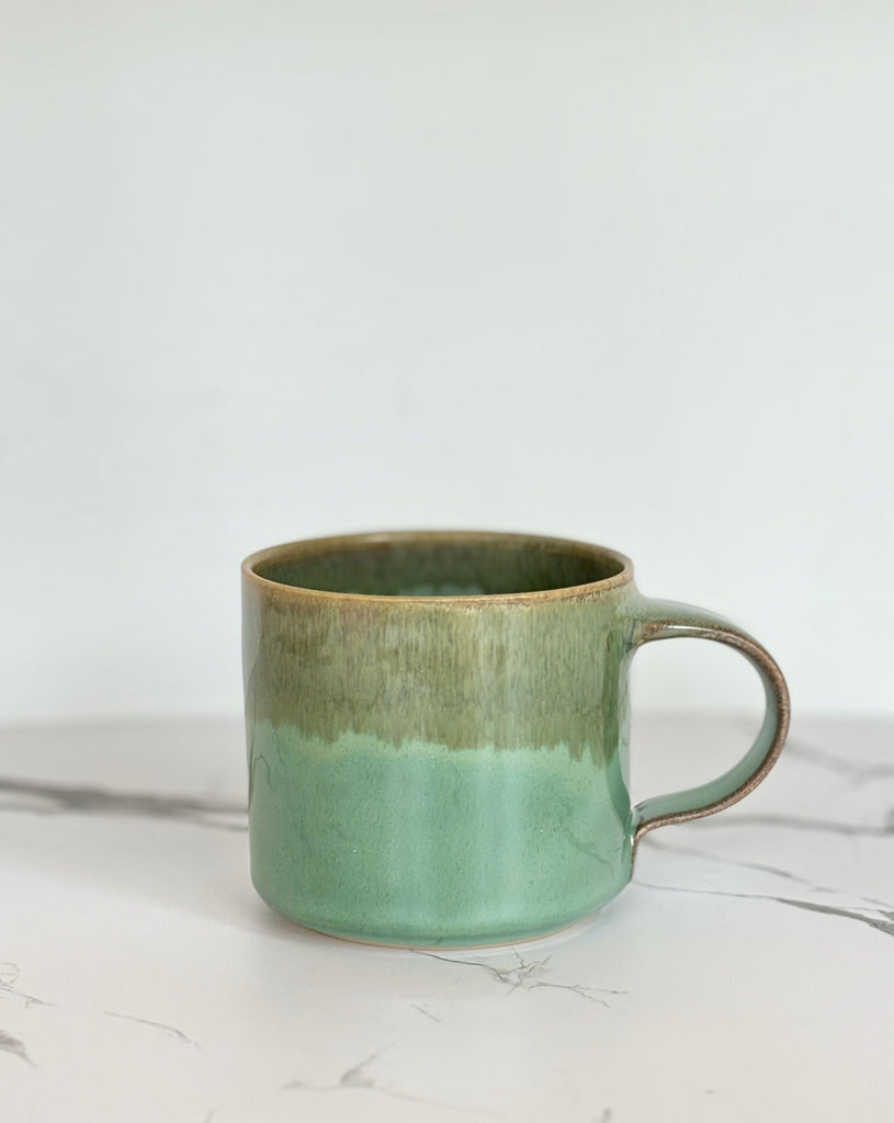 A green and brown two-toned gradient color ceramic mug with glossy finish on a marbled surface. 