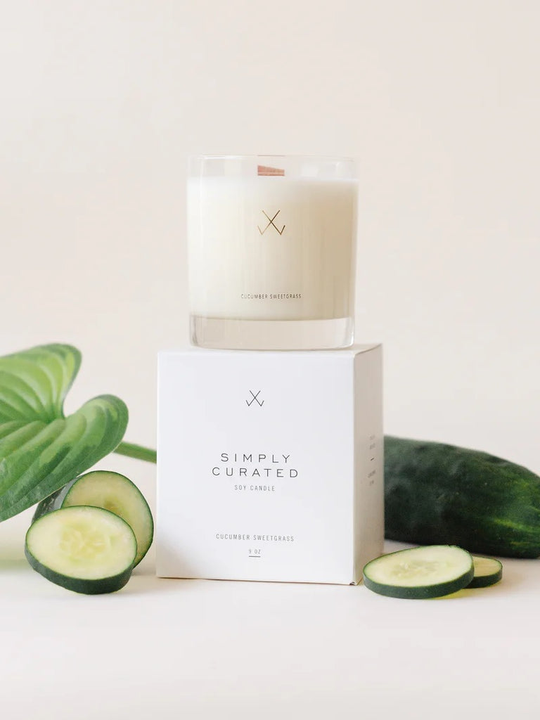 A Simply Curated cucumber sweetgrass soy candle on its white packaging box with minimalistic branding logo, with cucumber slices and a large green leaf on the side.